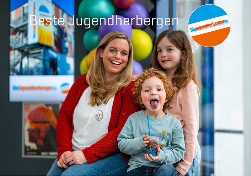 Beste Jugendherbergen. Eine fröhliche Familie, bestehend auf Mutter, Tochter und Sohn, in der Jugendherberge Trier. Die Mutter lacht, die Tochter lächelt und der Sohn hält einen Becher mit blauem Slush-Eis in der Hand und streckt die blaugefärbte Zuge raus. 